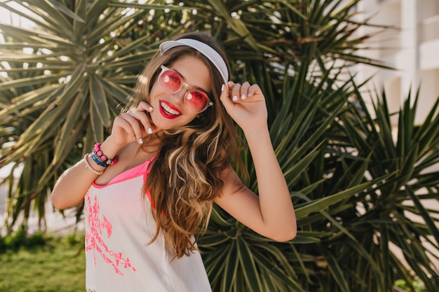 Joyful long-haired girl in trendy pink sunglasses gladly posing next to palm tree, enjoying vacation on exotic resort