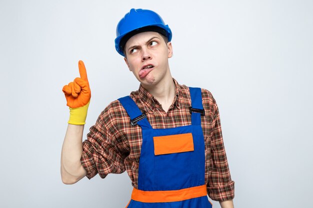 Joyful looking side showing tongue points at up young male builder wearing uniform with gloves 