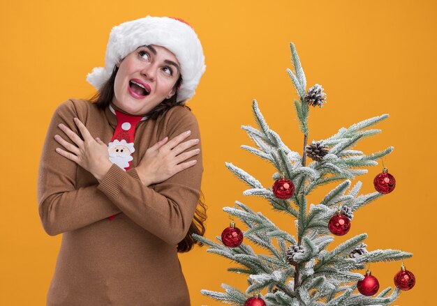 Joyful looking up young beautiful girl wearing christmas hat with tie standing nearby christmas tree putting hands on shoulder isolated on orange wall