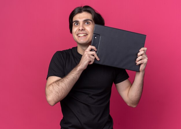 Joyful looking up young handsome guy wearing black t-shirt holding clipboard around face isolated on pink wall