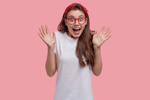 Joyful lovely girl has fun, gestures actively, laughs over something funny, wears red headband and casual t shirt, shows palms at camera