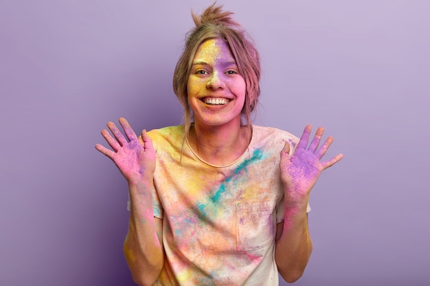 Joyful lovely young woman shows colorful palms, being in good mood after visiting Holi festival first, plays with colors, smeared with colored powder over purple wall