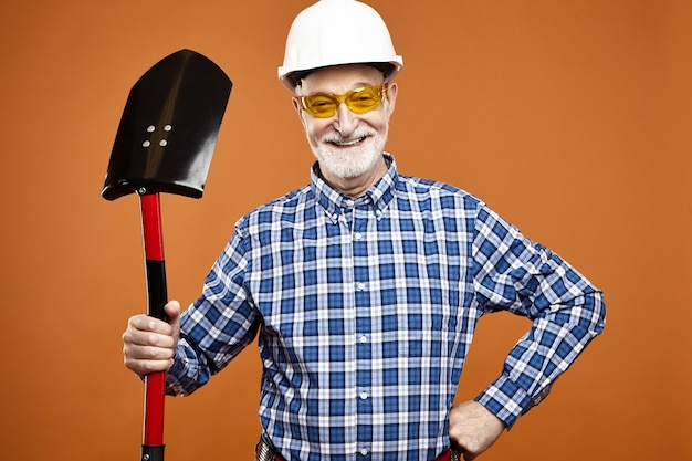 Foto gratuita gioioso impiegato edile maschio in pensione che indossa il casco protettivo e occhiali gialli, usando la pala per scavare, in posa isolato contro il muro vuoto del copyspace