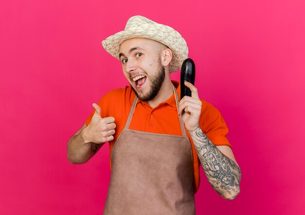 Joyful male gardener wearing gardening hat holds eggplant and thumbs up