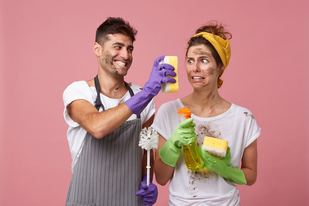 Joyful male wearing apron and protective gloves showing his wife dirty sponge very closely to her face presenting results of his work. dirty housemaid looking at dirty sponge with disgust or aversion