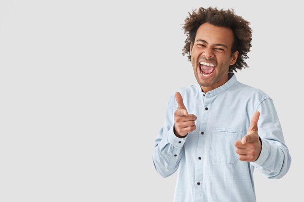 Радостный мужчина со счастливым выражением лица, широко открывает рот, у него вьющиеся волосы, показывает обоими указательными пальцами, делает выбор, одетый в белую рубашку, позирует над стеной, пустое пространство