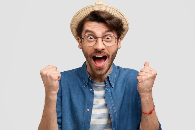 Joyful man agronomist clenches fists, opens mouth widely, exclaims with happiness, wears straw hat and casual denim shirt, isolated over white wall