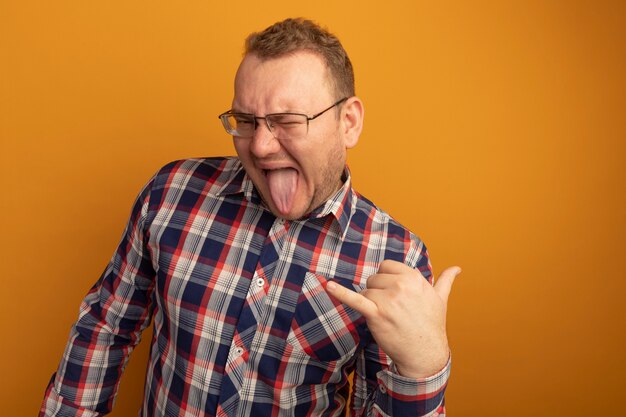 Joyful man in glasses and checked shirt happy and cheerful showing rock symbols sticking out tongue standing over orange wall