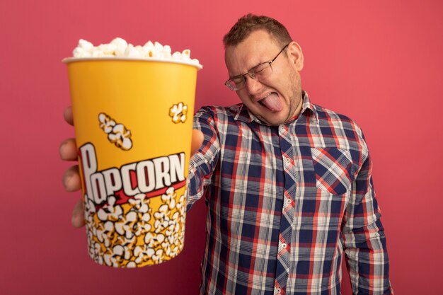 Joyful man in glasses and checked shirt showing bucket with popcorn sticking out tongue standing over pink wall