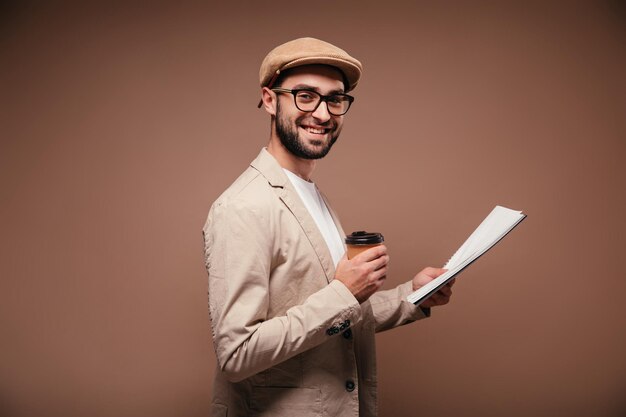 Joyful man in glasses looks into camera and holds notebook cup of coffee