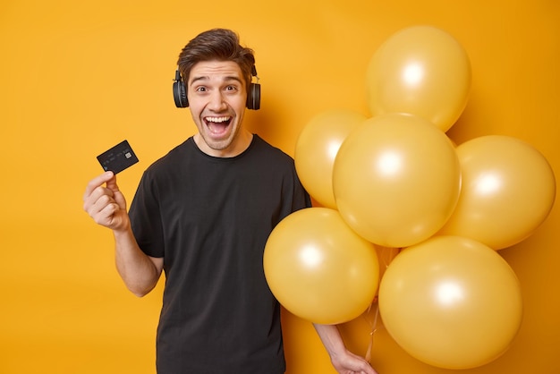 L'uomo gioioso felice di ottenere una grossa somma di denaro sul suo conto bancario pronto per festeggiare il compleanno tiene un mazzo di palloncini gonfiati e la carta di credito ascolta musica tramite le cuffie isolate sopra il muro giallo