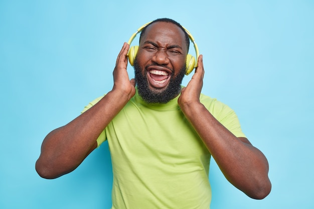 Joyful man with thick beard keeps hands on headphones laughs happily enjoys favorite music wears casual green t shirt isolated over blue wall