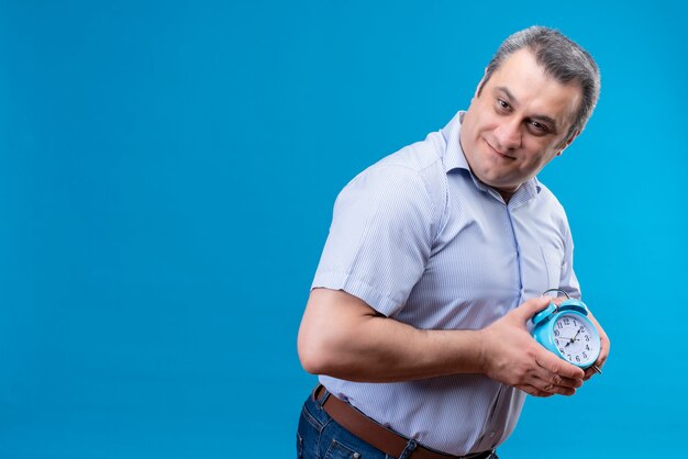 Joyful middle age man in blue striped shirt holding blue alarm clock with hands on a blue background