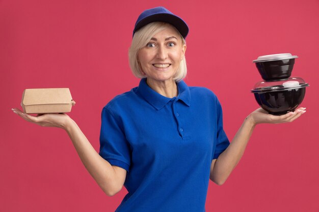 Joyful middle-aged blonde delivery woman in blue uniform and cap holding paper food package and food containers 