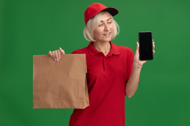 Joyful middle-aged blonde delivery woman in red uniform and cap holding paper package and mobile phone looking at phone