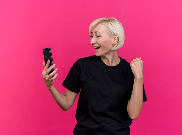 Радостная блондинка средних лет славянская женщина, держащая и смотрящая на мобильный телефон, делает жест да, изолированный на малиновом фоне