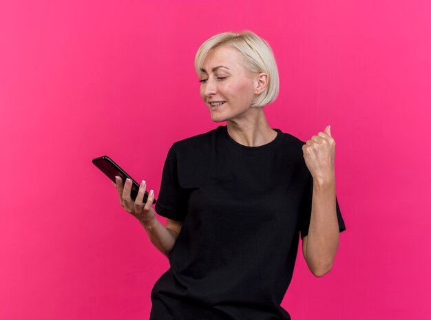 Радостная белокурая славянская женщина средних лет, держащая и смотрящая на мобильный телефон, делает жест `` да '' на малиновой стене