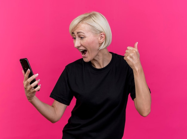 Радостная белокурая славянская женщина средних лет, держащая и смотрящая на мобильный телефон, показывает палец вверх изолированной на розовой стене