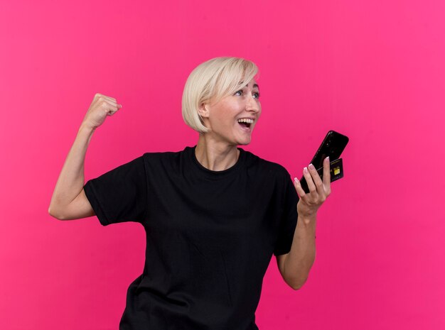 Joyful middle-aged blonde slavic woman holding mobile phone and credit card looking at side doing yes gesture isolated on crimson wall