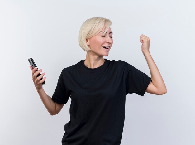 Joyful middle-aged blonde slavic woman holding mobile phone doing yes gesture with closed eyes isolated on white background