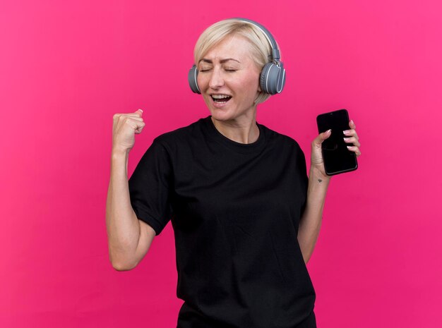 Joyful middle-aged blonde slavic woman wearing headphones holding mobile phone doing yes gesture isolated on pink wall