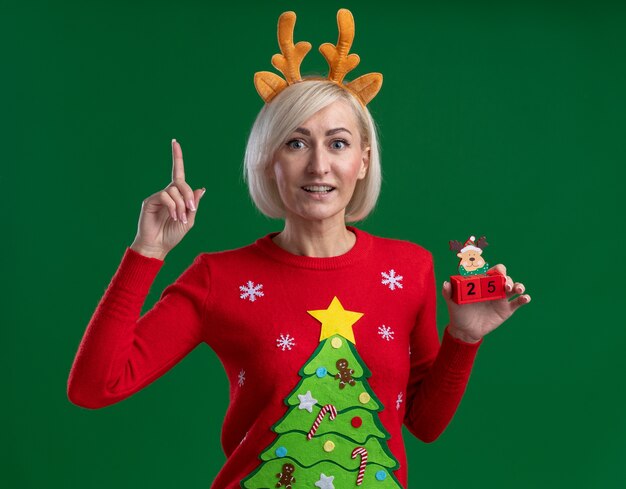 Joyful middle-aged blonde woman wearing christmas reindeer antlers headband and christmas sweater holding christmas reindeer toy with date looking at camera pointing up isolated on green background