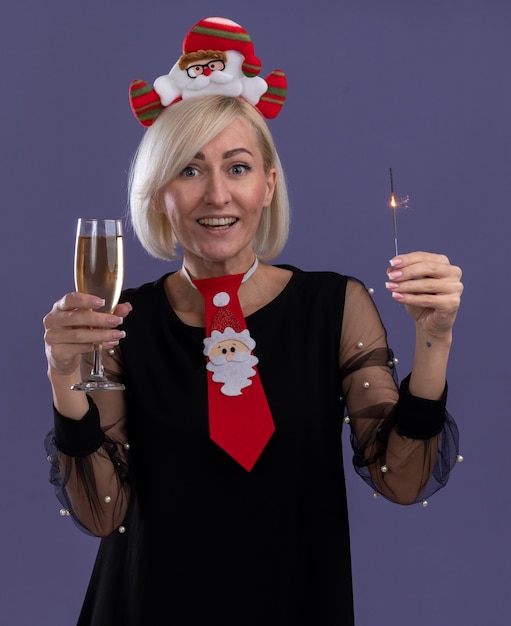 Gioiosa donna bionda di mezza età che indossa la fascia di babbo natale e cravatta holding sparkler vacanza e un bicchiere di champagne guardando la telecamera isolata su sfondo viola