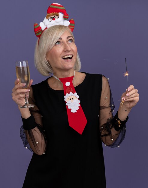 Joyful middle-aged blonde woman wearing santa claus headband and tie holding holiday sparkler and glass of champagne looking up isolated on purple background