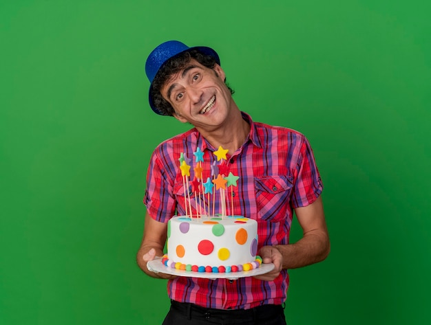 Joyful middle-aged caucasian party man wearing party hat holding birthday cake looking at camera isolated on green background with copy space