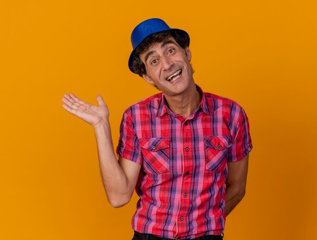 Joyful middle-aged caucasian party man wearing party hat looking at camera showing empty hand keeping another one behind back isolated on orange background with copy space