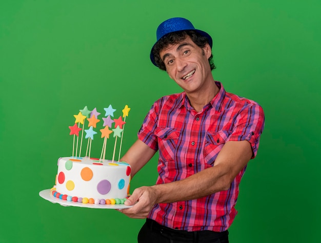 Joyful middle-aged caucasian party man wearing party hat stretching out birthday cake looking at camera isolated on green background