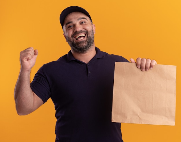 Радостный курьер среднего возраста в униформе и кепке держит бумажный пакет с едой, показывая жест да, изолированный на желтой стене