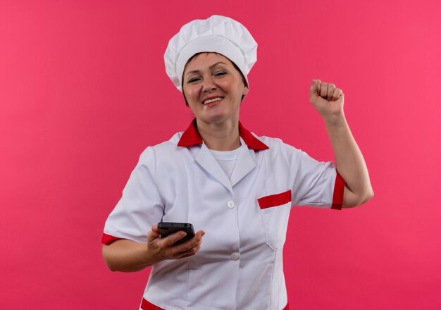 Радостная женщина-повар средних лет в униформе шеф-повара держит телефон и показывает жест да на изолированной розовой стене