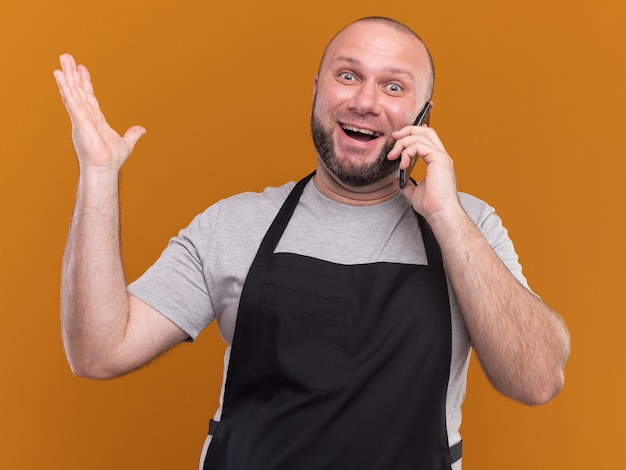 Joyful middle-aged male barber in uniform speaks on phone raising hand isolated on orange wall
