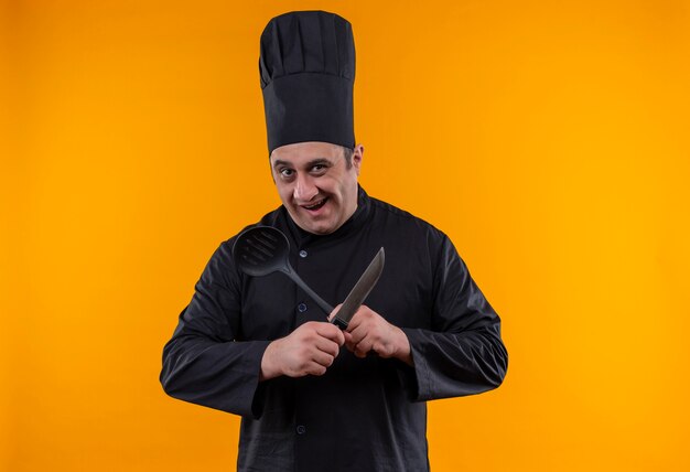 Joyful middle-aged male cook in chef uniform crossing knife and spatula on yellow wall