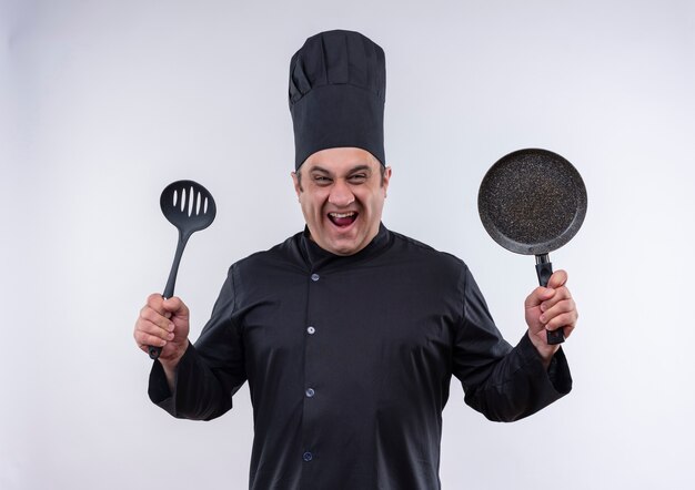 Joyful middle-aged male cook in chef uniform holding frying pan and spatula on isolated white wall