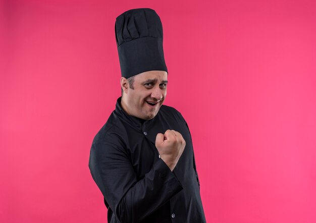 Joyful middle-aged male cook in chef uniform showing yes gesture on isolated pink wall with copy space