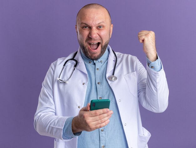 Joyful middle-aged male doctor wearing medical robe and stethoscope holding mobile phone doing yes gesture 