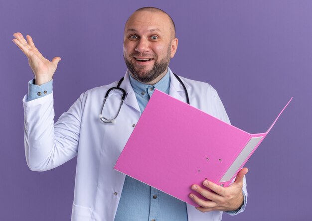Радостный мужчина-врач средних лет в медицинском халате и стетоскопе держит открытую папку, глядя вперед, показывая пустую руку, изолированную на фиолетовой стене