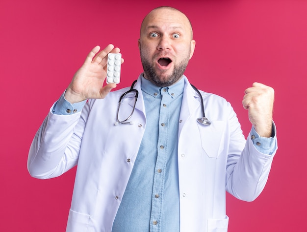 Free photo joyful middle-aged male doctor wearing medical robe and stethoscope showing pack of medical tablets to camera  doing yes gesture isolated on pink wall