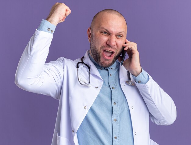 Joyful middle-aged male doctor wearing medical robe and stethoscope talking on phone looking at side doing yes gesture isolated on purple wall