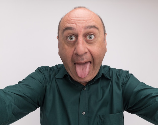 Free photo joyful middle-aged man wearing green t-shirt holding camera showing tongue isolated on white wall