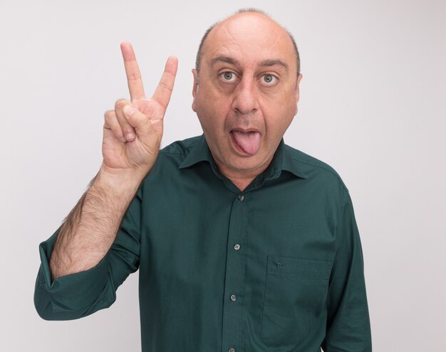 Joyful middle-aged man wearing green t-shirt showing tongue doing peace gesture isolated on white wall