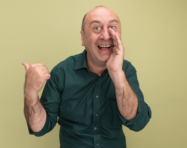 Joyful middle-aged man wearing green t-shirt whispers points at behind isolated on olive green wall with copy space