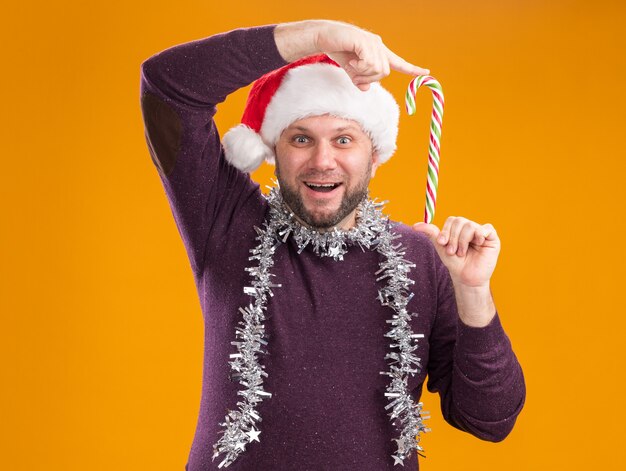 Joyful middle-aged man wearing santa hat and tinsel garland around neck holding christmas sweet cane near head  isolated on orange wall