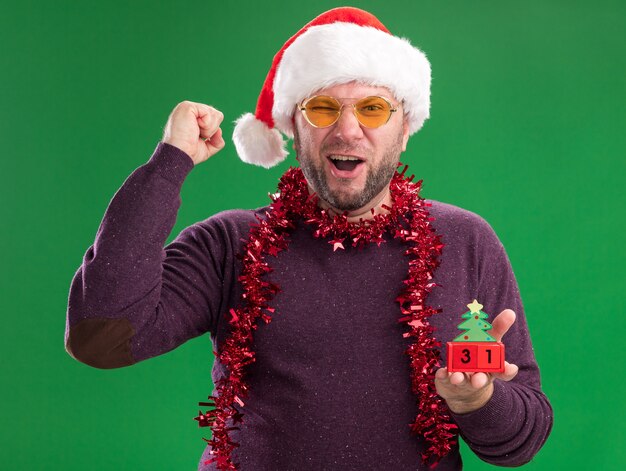 Joyful middle-aged man wearing santa hat and tinsel garland around neck with glasses holding christmas tree toy with date