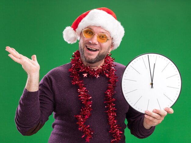 Радостный мужчина средних лет в шляпе санта-клауса и гирлянде из мишуры на шее в очках с часами, показывающими пустую руку, изолированную на зеленой стене