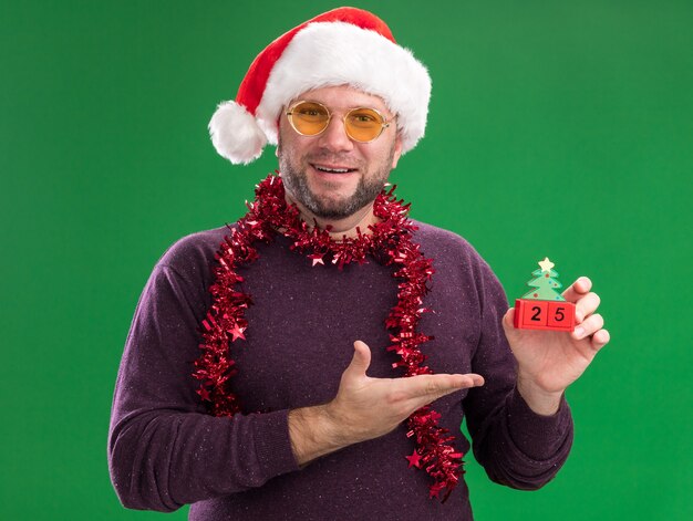 Радостный мужчина средних лет в новогодней шапке и мишурной гирлянде на шее в очках, держащий и указывающий рукой на елочную игрушку с датой, смотрящий в камеру, изолированную на зеленом фоне