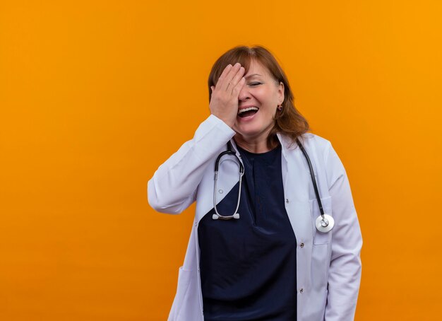 의료 가운 및 복사 공간이 격리 된 오렌지 벽에 눈에 손을 넣어 청진기를 입고 즐거운 중년 여성 의사