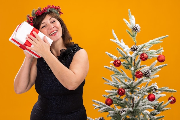 飾られたクリスマスツリーの近くに立っている首の周りにクリスマスの頭の花輪と見掛け倒しの花輪を身に着けているうれしそうな中年の女性